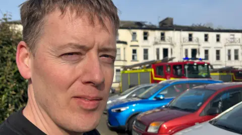 Andrew Turner/BBC A man with fair hire wearing a black t-shirt faces the camera with a fire engine and a fire-damaged building in the background.