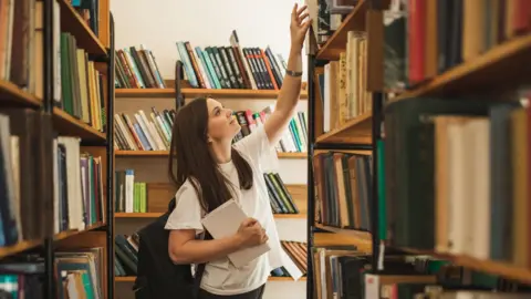 Getty Images A female student standing in university library. she is wearing a white t shirt and reaching up for a book on the top shelf.