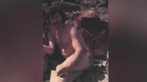 Family handout A man with black hair and a moustache sits on what appears to be a beach. He is topless and wearing swimming trunks.