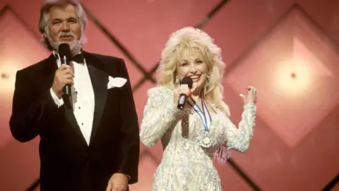 Beth Gwinn/Redferns/ Getty Kenny Rogers, wearing a black and white tuxedo, and Dolly Parton, wearing a bejewelled white and cream long sleeved dress, performing on stage with a pink background.
