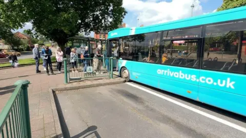 A single-decker turquoise bus parked in an open-air bus stand with a handful of passenger queuing to board. Green railing and a small bus stop can be seen alongside the bus.