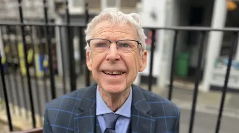 Deputy David De Lisle - a man with white hair smiling at the camera. He is wearing black top rimmer spectacles and a blue block checked suit with a blue shirt and a blue tie. 