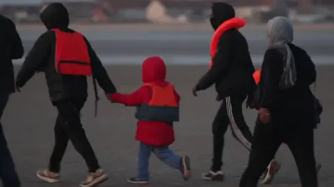 A group of four people, including what looks like two men, a woman in a hijab, and a child in a red coat, walking on a beach. They have orange lifejackets on.