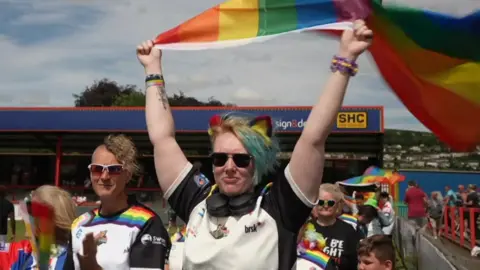 BBC A woman in a Keighley Cougars rugby shirt, with headphones resting around her neck and several colourful wristbands has both arms raised, holding a large rainbow Pride flag stretched overhead. To her left another supporter is visible wearing a similar Cougars shirt with rainbow detailing and sunglasses. 