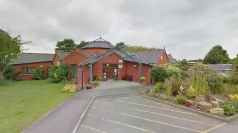 Google The Langport surgery entrance surrounded by flowerbeds, with a large glass tunnel leading to the front door