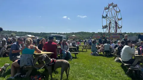 Crowds of people eating on picnic tables and enjoying the Sherborne Castle Country Fair in the sun. There are some dogs as well. There is a Ferris Wheel in the distance and a white ice cream van nearby.