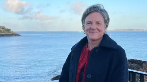 Deputy Sally Rochester is standing in front of a view of Guernsey harbour, with the sea behind. She has short grey hair and is wearing a blue wool coat. She is smiling at the camera.