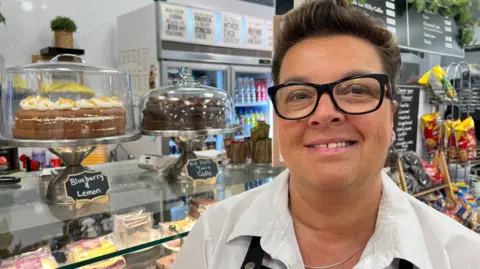 A woman smiles as she stands in a cafe in front of a counter piled with cakes and other food. She has dark-brown short hair, swept back, and wears black-rimmed glasses, a white shirt and a black apron. Fridges and black menu boards are in the background.