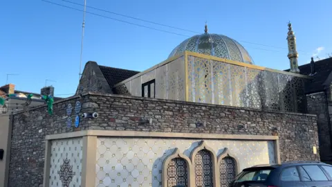 An exterior view of Easton Jamia Masjid. The building has an exterior brick wall and the internal section of the mosque has patterned windows and an ornate dome.