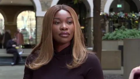 A woman with light brown hair and a black top looks slightly off camera, with the background blurred. 