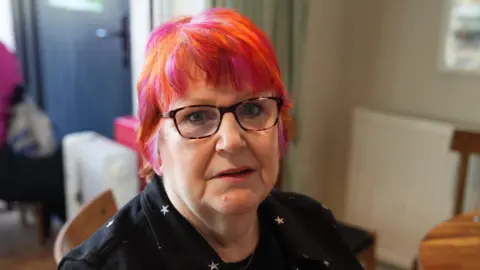 Richard Knights/BBC Avril Green wears a black shirt with silver stars. She has tortoiseshell glasses and cropped hair which is pink and orange.