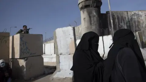 EPA An Israeli soldier watches as Palestinians women wait to cross Qalandia checkpoint to attend the first Friday prayer service of Ramadan at the al-Aqsa Mosque in occupied East Jerusalem (16 April 2021)