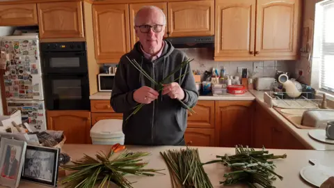 Sean Lipfird Sean Lipfird making St Brigid's crosses in his kitchen in Kircubbin.  He has short, grey hair and glasses and is wearing a grey hooded jacket over a pink shirt. 
He is holding rushes and there are piles of rushes on the counter top. 