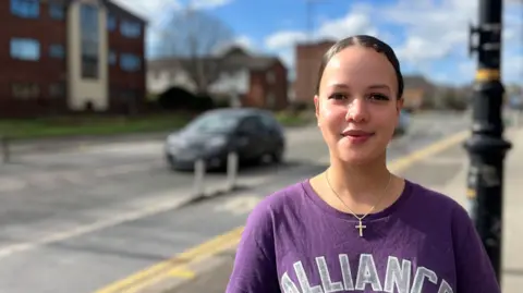 BBC Amelia with brown hair tied back wearing a purple t-shirt with a silver cross in a street near Old Trafford