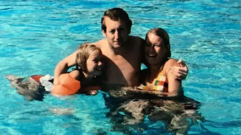 Handout A family of three comprising a young girl, man and woman smile as they have their arms around each other in a swimming pool