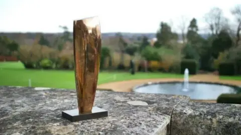 Bryanston School A plain polished bronze sculpture on a stand. It appears to be the size of a forearm and has been placed on a stone-capped wall in the open air. In the background is a lawn with a round pond and fountain.