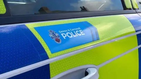 A close up shot of the outside door of a Devon & Cornwall Police car. The forces branding is on the outside in a blue rectangle. The regular police colours of yellow and blue are wrapped around the vehicle.

