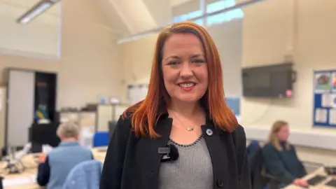 Nicola is standing behind a office with people working blurred in the background. She is wearing a grey top with black beads on the collar and a black jacket.