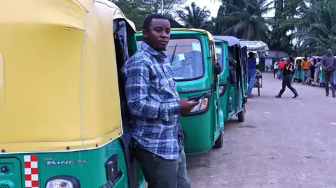 BBC A man in jeans, sliders and a checked shirt stands by his three-wheeled vehicle as he waits to fill up with CNG.