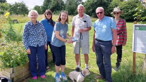 Devon Wildlife Trust Five members of the Friends of Downhorn Park, one with a small dog on a lead, receive their award from Devon Wildlife Trust's Hetty Lewis. The group are standing in the garden in the sunshine