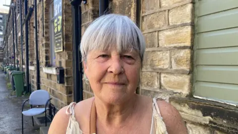 A woman with a grey bob hairstyle standing outside next to a building made of yellow Yorkshire stone.