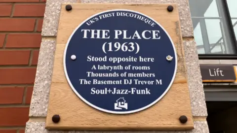 A large blue plaque on a wooden board says 'The Place, 1963, stood opposite here. 'A labryinth of rooms, thousands of members. The Basement DJ Trevor M, Soul and Jazz Funk'