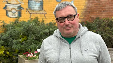A headshot of Calne town councillor Michael Hudston wearing black glasses and a grey zip-up hoodie, standing in front of some plants, flowers and a yellow-painted brick wall. 