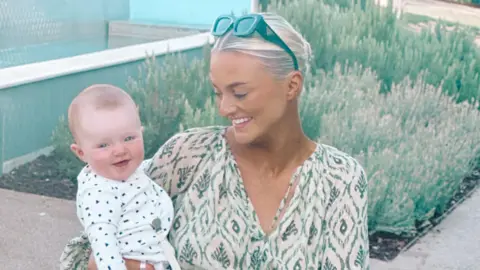 Kelly Medina Enos Kelly Medina Enos holding her daughter in her arms and smiling at her. She is in front of some plants