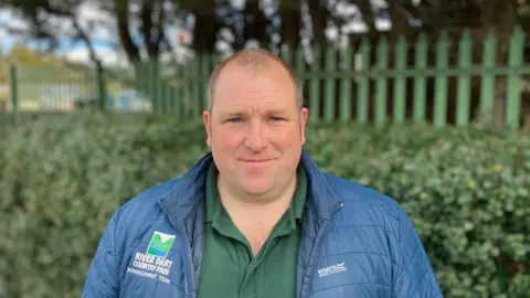 A man in a blue jacket stands in front of a hedge at Crealy Theme Park in Devon. It says River Dart Country Park on his coat. 