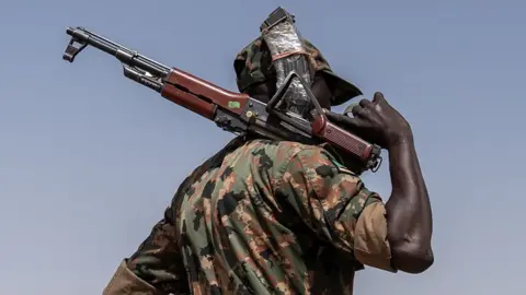 Avaaz via Getty Images A metade superior de um homem em um uniforme militar contra um céu azul. Ele é afastado da câmera e está segurando um rifle automático por cima do ombro.