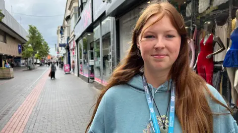 Katie, 17, in Middlesbrough. She has auburn hair and a light blue hoodie. She wears a college lanyard.
