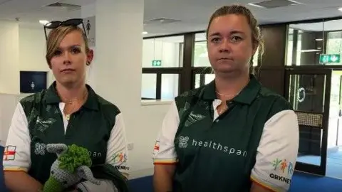 Zoe and Lisa stand next to each other. One is holding a cuddly toy mascot. They are wearing their Island Games kit, which is green and white.