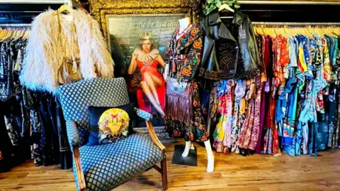 Want That Dress An inside view of Want That Dress shop with rails of colourful clothing and a chair and several jackets on hangers.