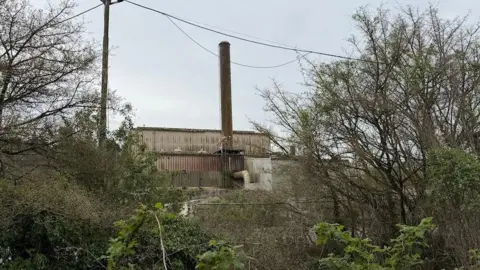 BBC An abanoned building in the middle distance with a large chimney coming out the top. It's built with corrugated metal. In the forground there are security fences and trees.