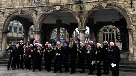 Peterborough City Council A choir dressed in black and wearing red Santa hats and white ribbons singing in front of the arches of a guildhall 