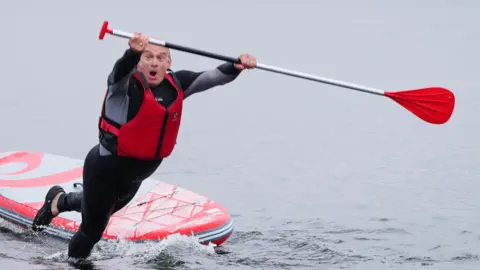 PA Media Ed Davey fakes surprise as he falls into the water from a paddleboard, wearing a black wetsuit and red life preserved and holding a paddle