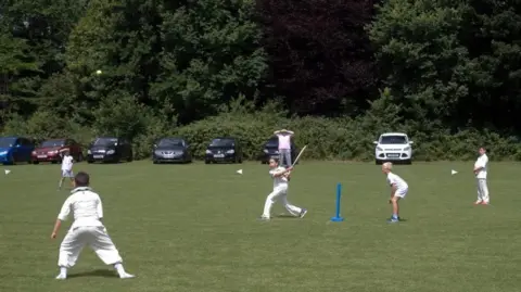 Libby Thomas Libby batting amongs a ring of four fielders. She is in her stance in front of free-standing blue plastic stumps. The cars parked in the background look as though they may be in danger