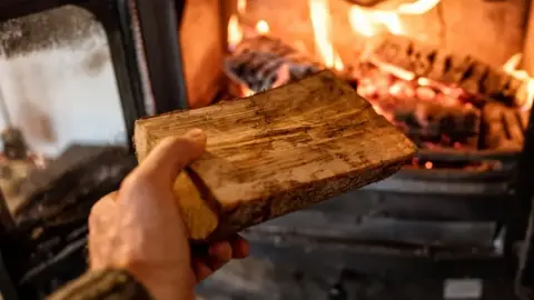 a hand holding a piece of wood that is about to be place into an already burning stove