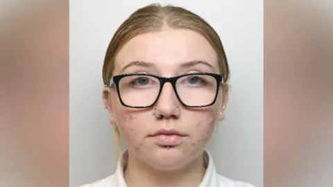 A woman with black-framed glasses, two nose piercings and a lip piercing looks at the camera.