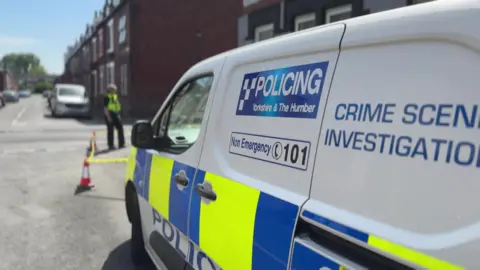 BBC A police Crime Scene Investigation van in the foreground, parked on a residential street. In the background, a police officer can be seen stranding on the street, with cars in the distance.