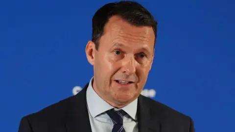 Russell Findlay wears a black blazer and grey shirt and is standing in front of a blue backdrop