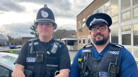 PC Davies and PCSO Newborough stood side by side and looking into the camera. Davies, stood on the left, is wearing a black police helmet with a silver crest on it, a black short sleeved shirt, and a black vest with a bodycamera and radio attached to it. On the vest is written 'Police' in white letters on a blue label. Newborough, on the right, is wearing an identical vest with the same equipment, with a blue short sleeved shirt underneath and a peaked police hat. They are stood in front of a row of police cars, outside a two storey brick building. The sky is cloudy and the sun is starting to set. 