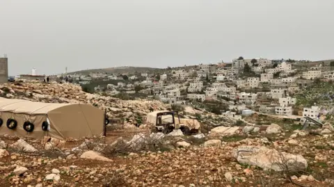 A view of Al-Mughayyir. Dozens of white breeze block homes dot a hillside covered in brown craggy rocks and green grass