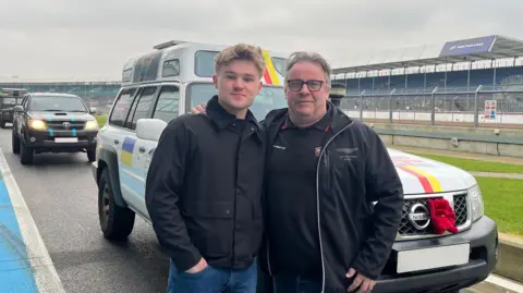 A young blonde man wearing a black coat and his dad, a man with glasses and grey hair. They've got their arms round each other and are smiling at the camera with a white Nissan vehicle behind them.