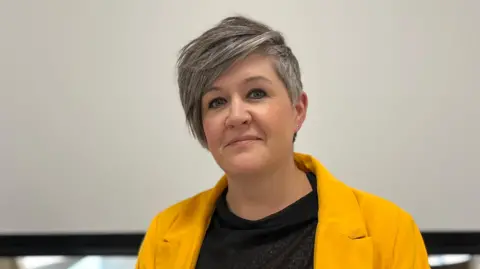 Deputy Haley Camp - A woman with black and grey hair, wearing a yellow jacket, and a black blouse. 