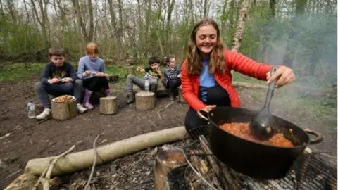 Children sitting outdoors in woodland. They are earing a meal on paper plates. A girl near the camera is stirring a pot of stew over a fire.