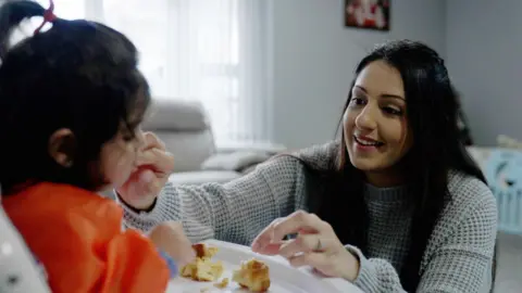 Harleen, a tall, dark-haired woman wearing a light-coloured jumper and thick weave, is smiling as she feeds her daughter in her high chair. Her daughter, wearing bright orange, is sitting with a few pieces of bread or pastry on the table in front, licking the pieces from her mother's fingers.