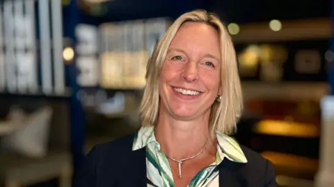Martin Giles/BBC Jenny Hanlon is sitting in the bar area of the Swan Hotel. There are table and chairs ready for customers in the background. Jenny is wearing a green and cream dress with a navy jacket and silver necklace. She is smiling at the camera.