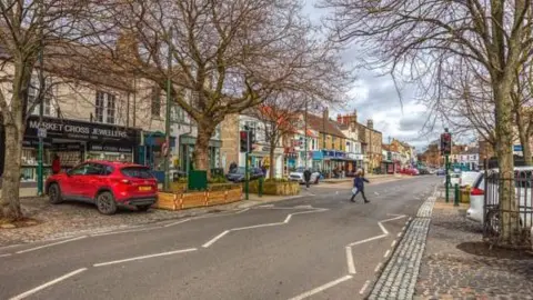 Lichfields Westgate in Guisborough. Cobbles line the road, while tall trees are growing  in planters along the road. A jewellers is among a number of shops on the high street.