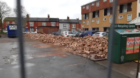 Leicester City Council A large pile of rubble, formerly garages in front of a row of cars. There are flats and houses in the background